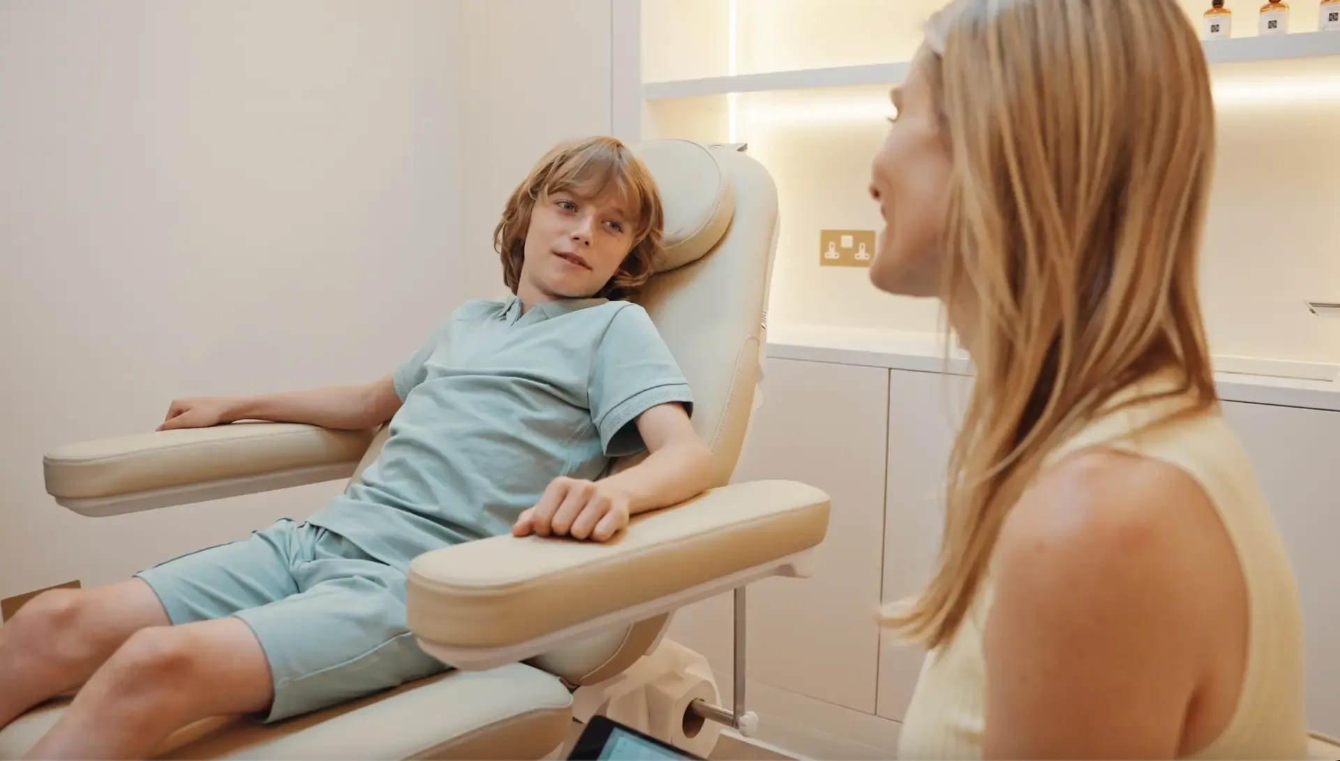 A young boy with brown hair in a blue hospital gown sitting in a beige examination chair, talking to a woman with long blonde hair in a cream-colored sleeveless top in a medical office.